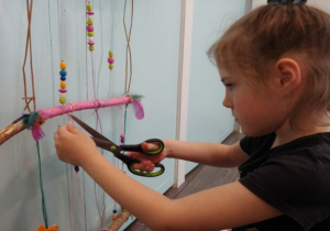 dziewczynka przycina nożyczkami sznurki, gdy robi instalację na ścianę ze sznurków, patyków, koralików
